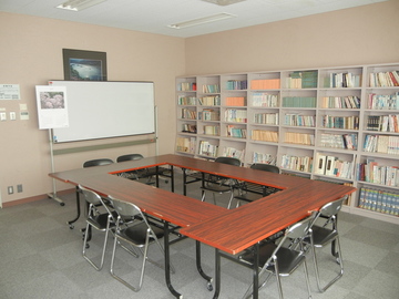 ロ状にテーブルが設置されており端の本棚に本が並べられている室内の写真
