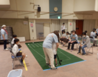 周囲で人々が見守る中、ボールの前で棒を下ろしている子供とサポートしている女性の写真