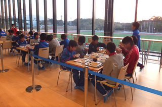 窓際のテーブル席に着いて机の上に用意された食事を大勢で食べている子供たちの写真