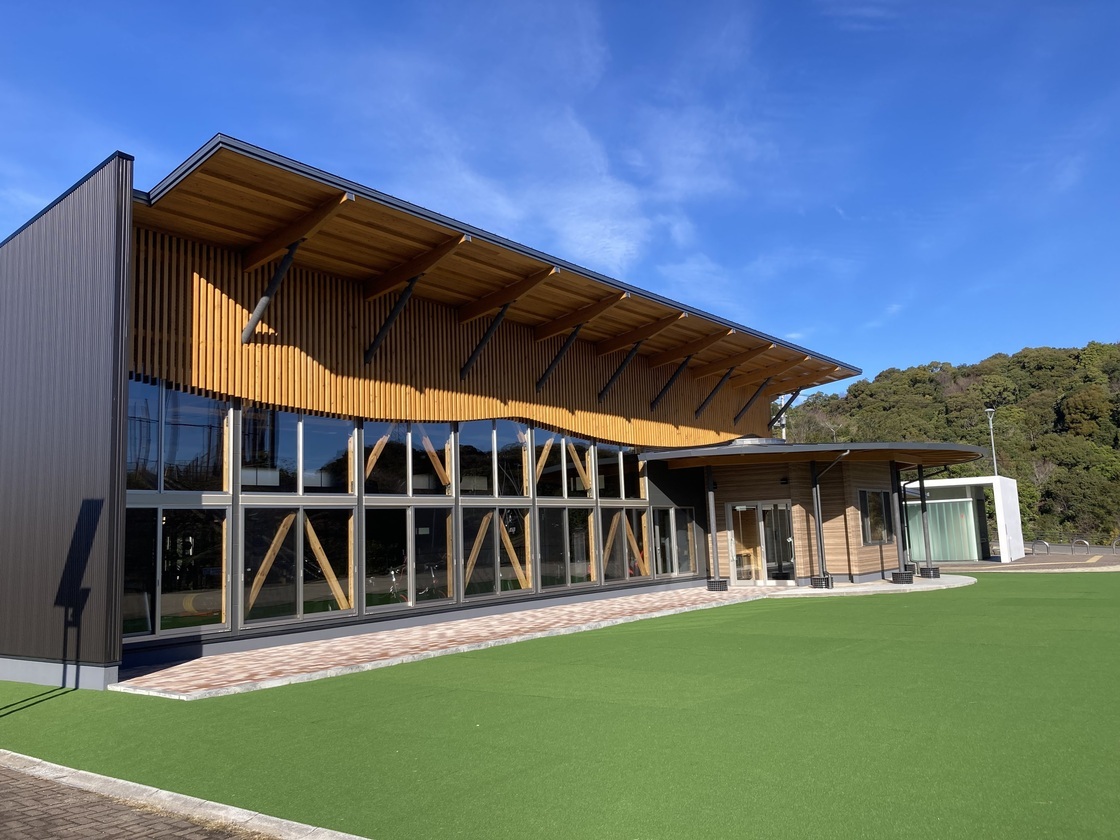 芝生のグラウンドに囲まれた木張りの屋根で奥行きがある建物の外観写真