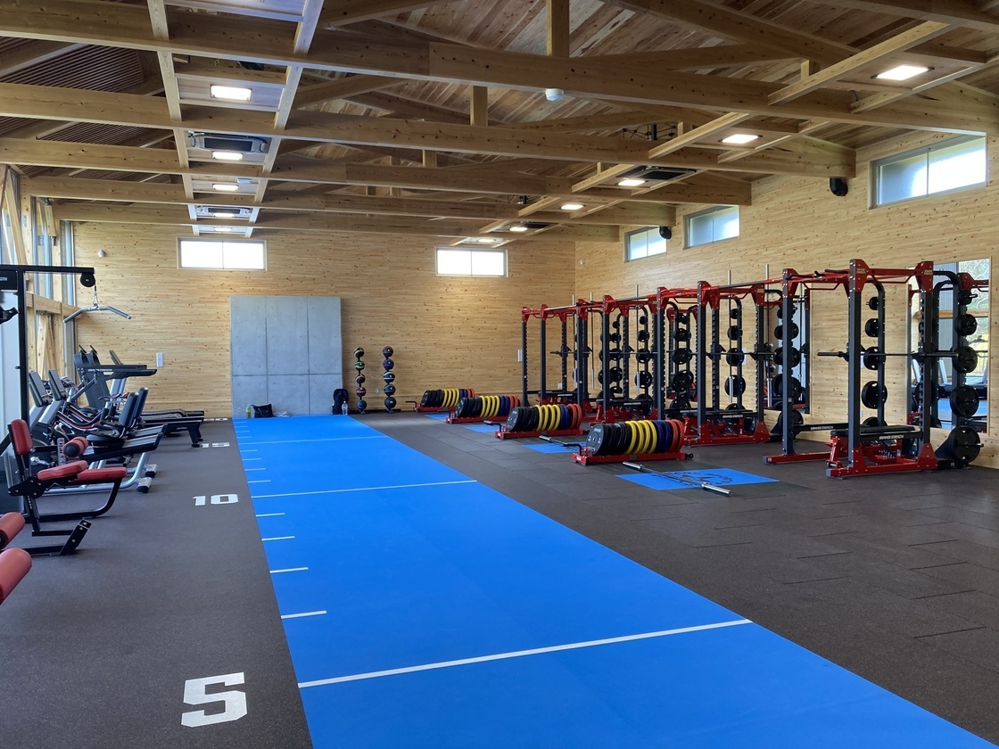 部屋の両側にウェイトトレーニングのマシンが何台も設置されている木張りの屋根をした建物の屋内写真