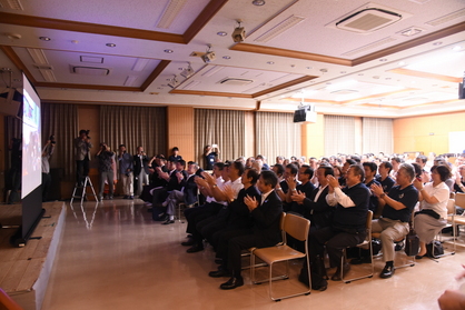 スクリーンの前で椅子に座っている多くの人々が拍手している写真