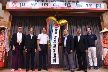 割られたくす玉から出る「世界遺産追加登録」と書かれた垂れ幕の両隣に男女が並んでいる写真