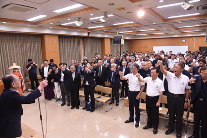 壇上の男性に向かって立ち上がった人々が腕をあげて乾杯している写真