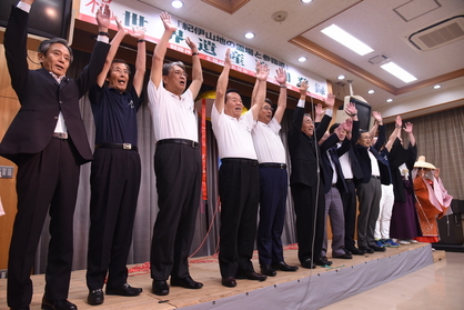 壇上で男性たちが両手を上げて万歳をしている写真