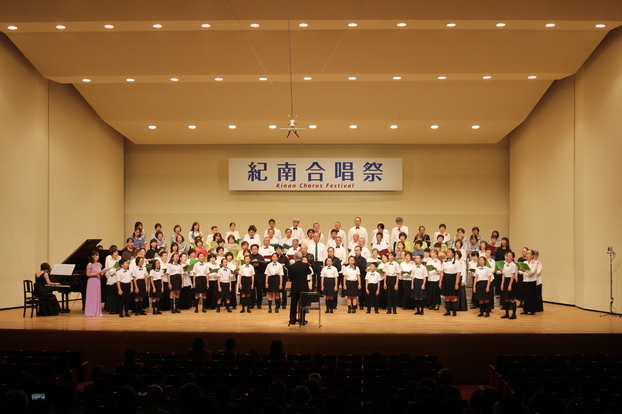 舞台で大勢の人が指揮者の前に並んで歌っている写真