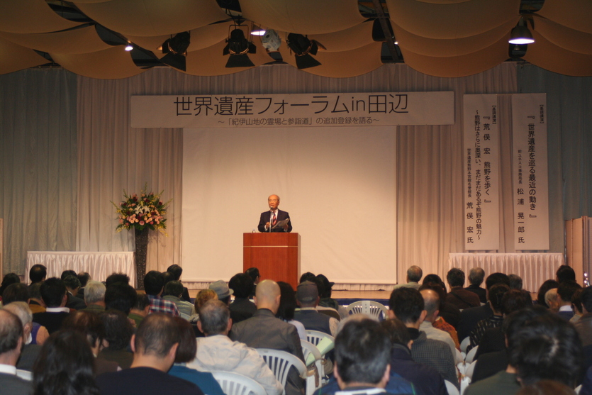 壇上でスーツを着た男性が大勢の観客の前でマイクを使って喋っている写真