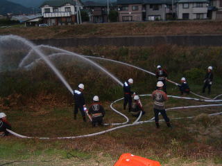 河川敷で制服を着た団員たちが斜面に向けて一斉に放水している写真