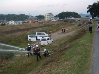 河川敷で制服を着た団員たちが斜面に向けて放水している全体の写真
