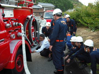 消防車の前で制服を着た団員たちが資機材の説明を聞いている写真