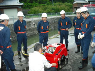 可搬ポンプの前で制服を着た団員たちが操作説明を受けている写真