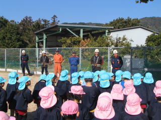 園庭で園児たちが整列した消防団員の話を聞いている後ろ姿の写真