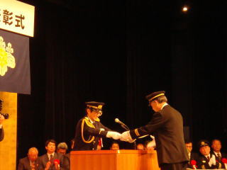 式典会場の壇上で消防団員が表彰状を受け取っている正面からの写真