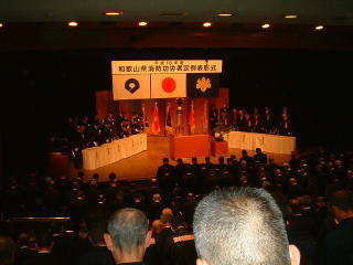 式典会場で消防団員や来賓が着席して開会式に臨んでいる後ろ姿の写真