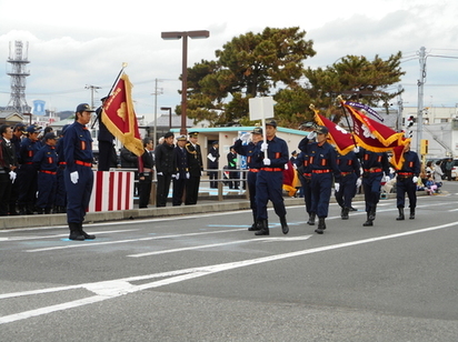 沿道で消防団員たちが団旗を掲げて行進している正面からの写真