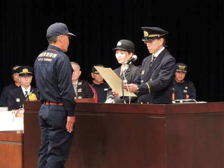 式典会場の壇上で消防団員が表彰状を受け取っている正面からの写真