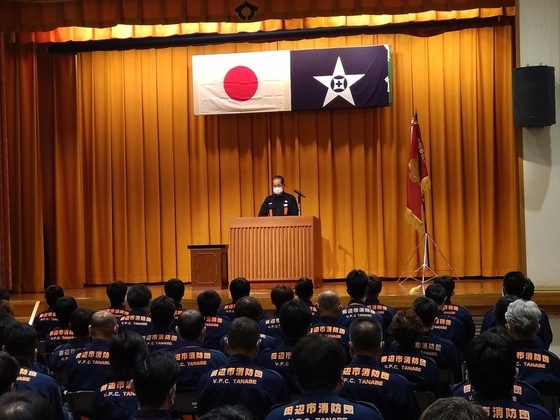 講堂の舞台で制服姿の団員に団長が訓示を述べている様子の写真