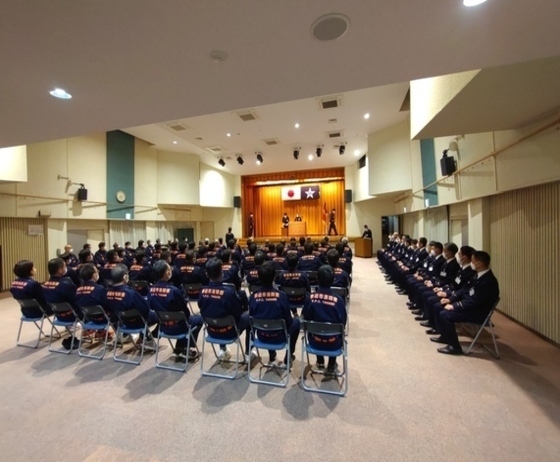 制服姿の団員たちが整列して座り、式典に参加している様子を写した体育館の後方からの写真