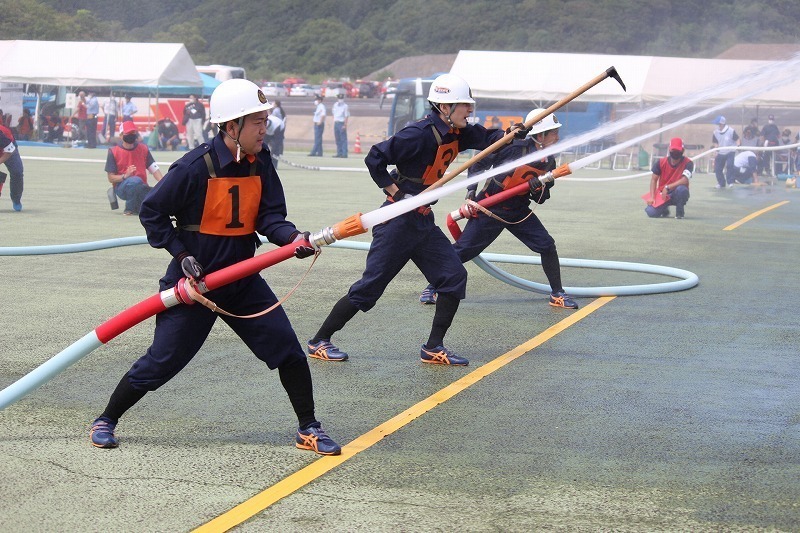 運動場で制服を着た団員たちがホースを構えて一斉に放水している写真