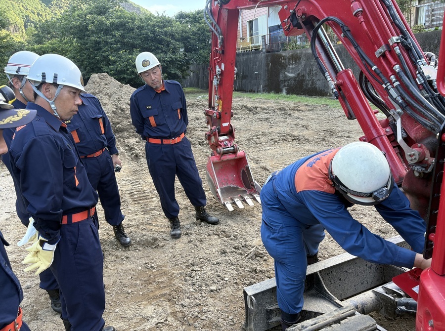 防火服を着た隊員が、赤いショベルカーのショベル根本部分を操作している様子を見守る防火服を着た5人の隊員の写真