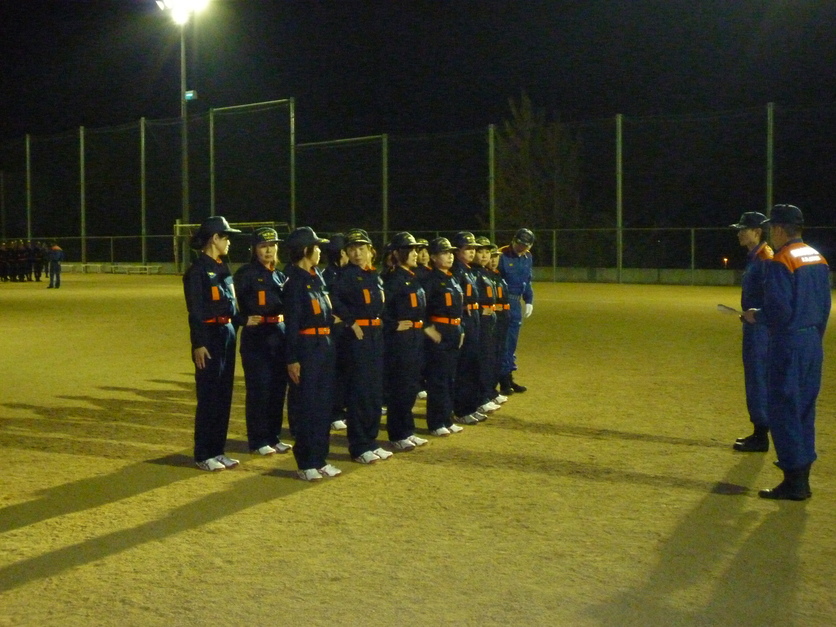 夜の運動場で防火服を着た団員たちが整列して点呼を受けている写真