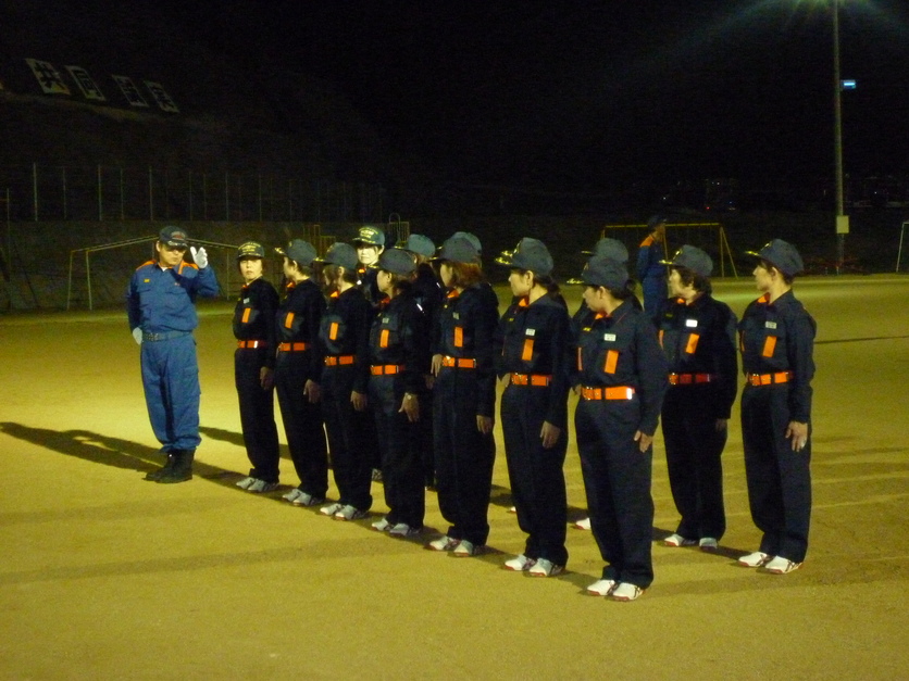 夜の運動場で防火服を着た団員たちが教官に向かって整列している写真