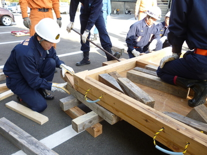 駐車場で防災服を着た職員たちが木材を使って簡易構造物を組み立てている写真