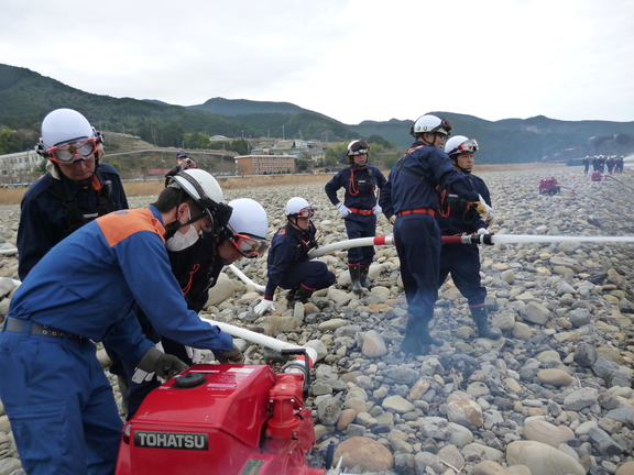 河原で制服を着た団員たちが可搬ポンプを操作して放水訓練をしている写真