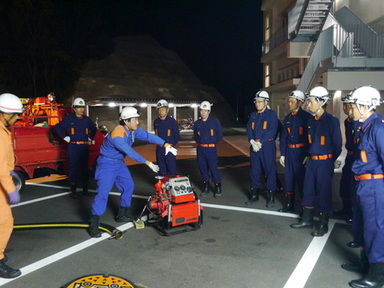駐車場で防災服を着た職員たちが発電機の操作方法を実演で学んでいる写真