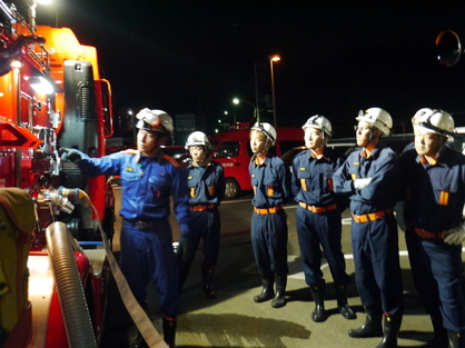 消防車の横で防災服を着た職員たちが放水機材の説明を受けている写真