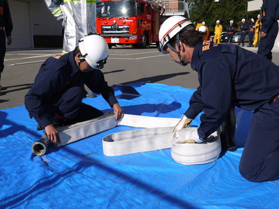 ブルーシートの上で防災服を着た職員たちがホースを折りたたんで整えている写真