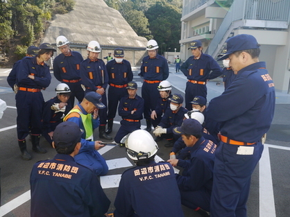 建物の前で防災服を着た職員たちが円になって訓練の振り返りをしている写真