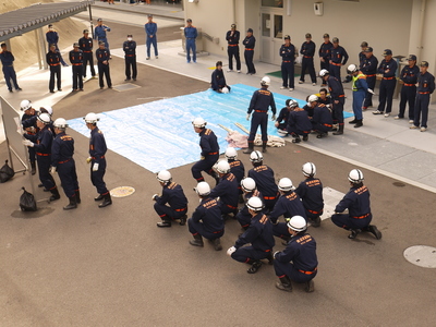建物の前で防災服を着た職員たちがブルーシートを囲んで訓練の確認をしている写真