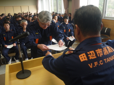 講堂に設置された演台に立った職員から防災服を着た職員が修了証を受け取っている写真