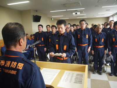 講堂に設置された演台に立った職員へ防災服を着た職員が挨拶を行っている写真