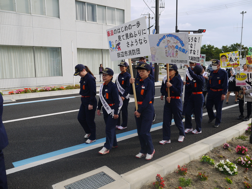 市街地の道路で消防団員たちがプラカードを掲げてパレードをしている写真