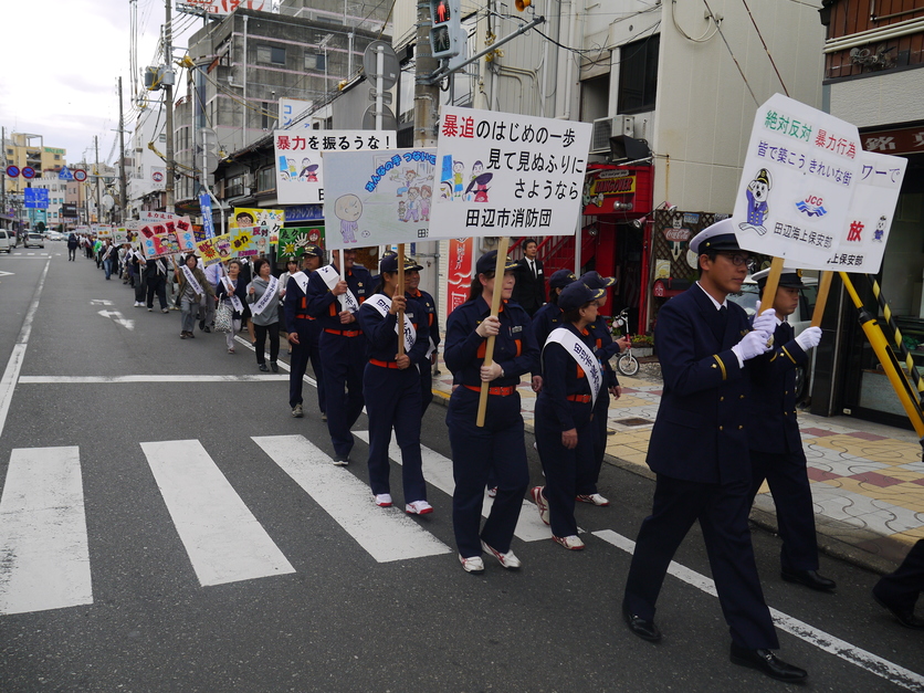 市街地の道路脇を整列して歩き、消防団員たちがプラカードを掲げてパレードをしている写真