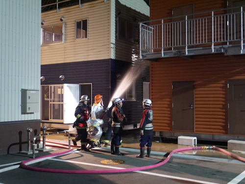 建物の前で防火服を着た団員たちがホースを構えて放水訓練をしている写真