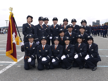 屋外会場で制服姿の消防団員たちが団旗を囲んで記念撮影している写真
