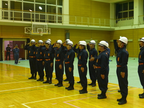 体育館で消防団の制服を着た消防団の団員たちが整列して敬礼している写真