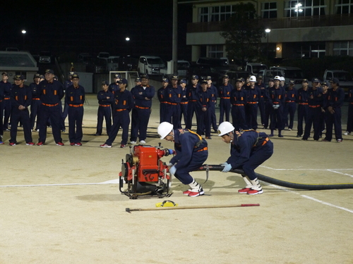 運動場で消防団の制服を着た団員たちが可搬ポンプを操作して放水準備をしている写真