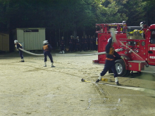 運動場で消防団の制服を着た団員たちがホースを操作して放水訓練をしている写真