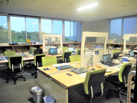 部屋全体を囲むような大きな窓が特徴的なパソコンが並んだ職員用デスクの全体を写した写真