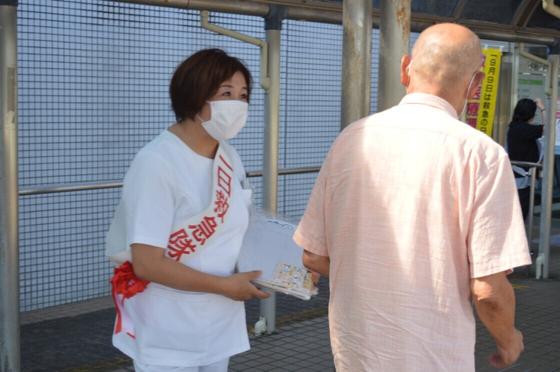 白地に赤字で「一日救急隊」と書かれたタスキをかけながら街頭に立っている女性の写真