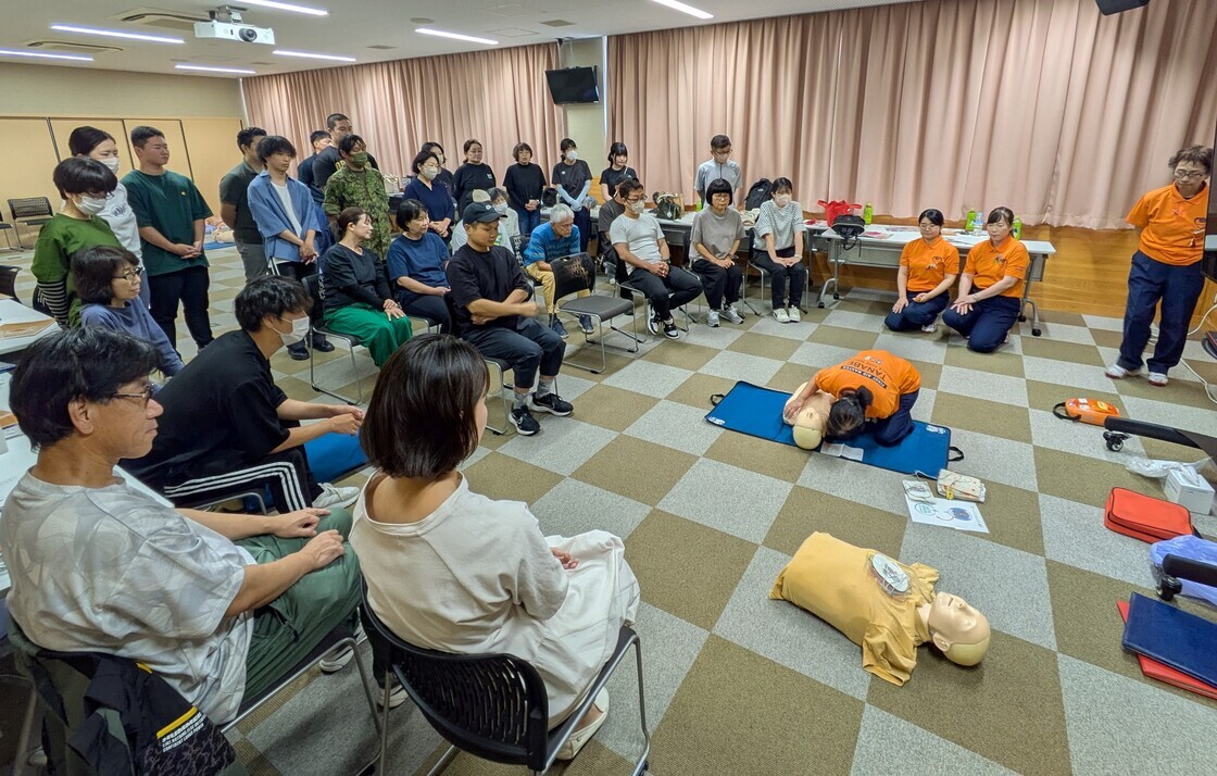 マットの上に横に置かれた人体の上半身の模型に向かってしゃがみこんで措置を施しているオレンジのTシャツを着た女性と、それを見ている大勢の人々の写真