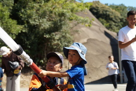 帽子をかぶった子どもが男性と一緒にホースを手に持ち放水している写真