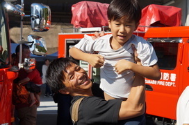 消防車の前で男の子が大人の男性に脇の下をつかんで持ち上げられている写真