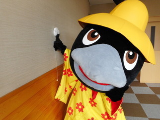 壁に設置された火災報知器の点検ボタンを押すカラスのキャラクター着ぐるみを着た人物の写真