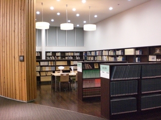 手前に仕切りがあり、大型の書籍が蔵書されている本棚と机といすが並んでいるエリア全体の写真