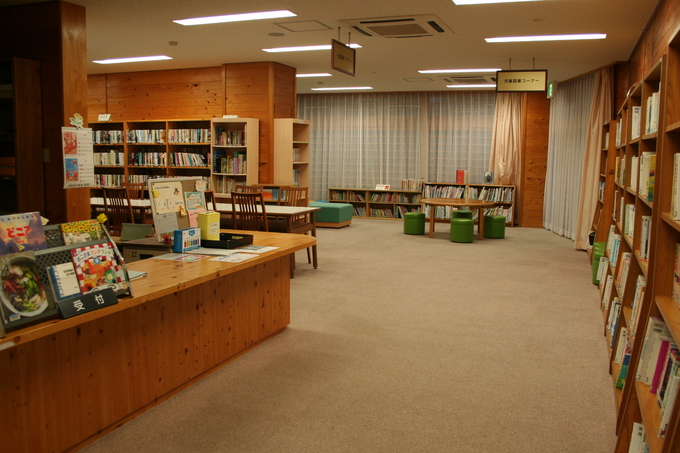 木の本棚やカウンターやテーブルがあり、緑系の椅子がある図書館の室内の写真